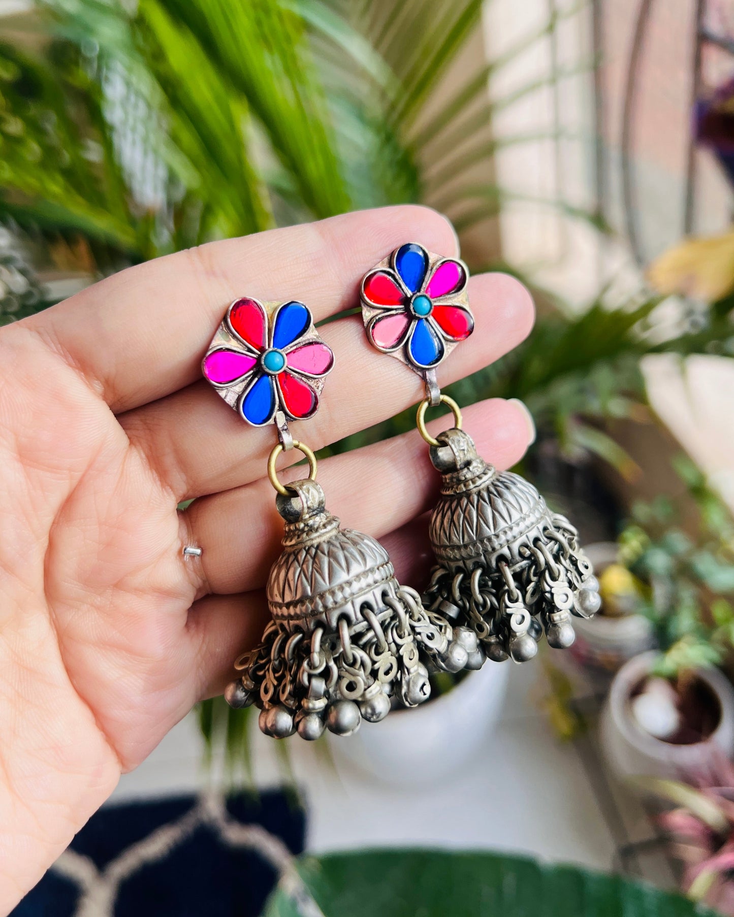 Colorful flower-shaped earrings with silver jhumkas held by a hand against a blurred natural background.