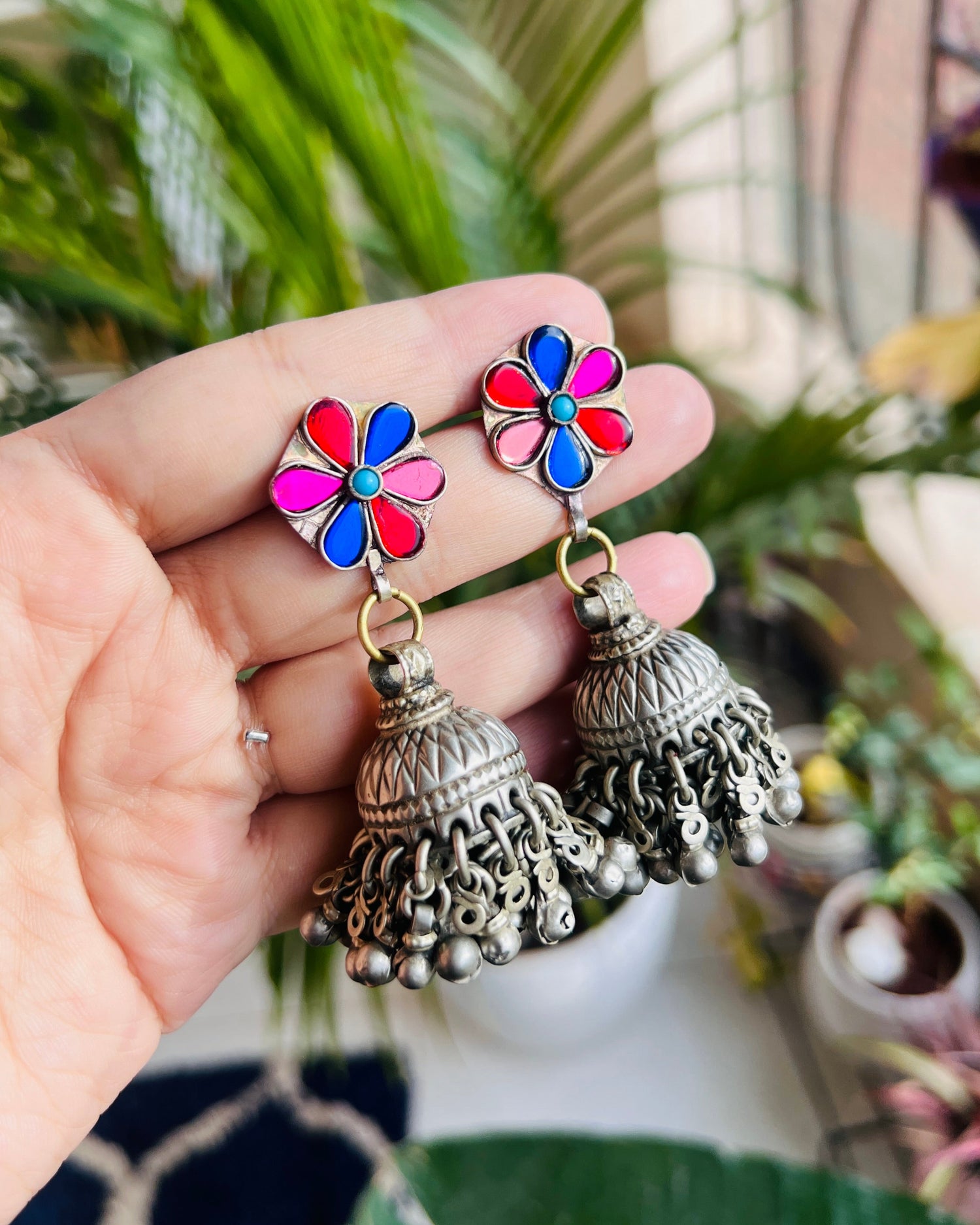 Colorful flower-shaped earrings with silver jhumkas held by a hand against a blurred natural background.